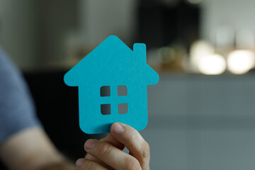 Close-up of a hand holding a small blue paper house cutout.  Home ownership, real estate, and new...