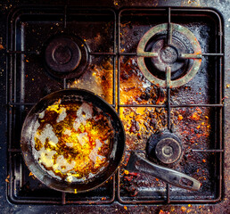 Dirty stove with a dirty black frying pan with leftover fried food. Unclean gas kitchen cooktop with greasy spots, old fat stains, fry spots and oil splatters.