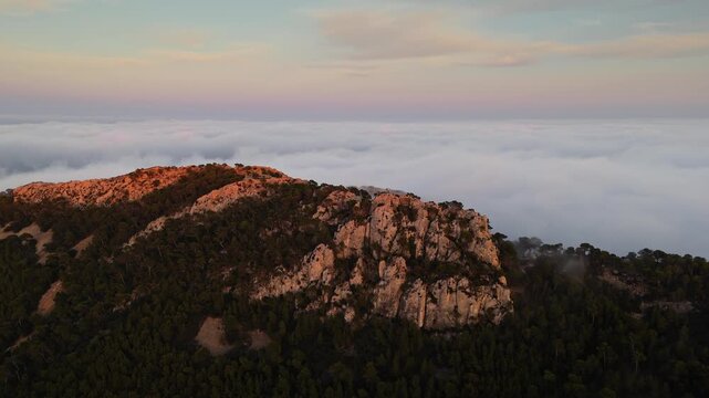Aerial drone video of a forested mountain peak in Spain, bathed in warm sunlight, emerging above dense clouds at sunrise.