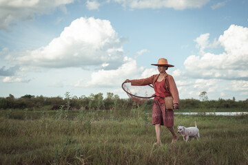 Serene rural landscape with farmer and dog carrying fishing net under a blue sky with fluffy clouds in a green field by the riverbank