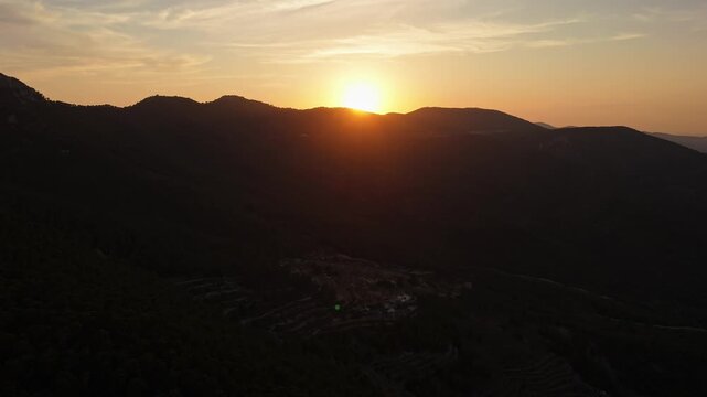 Aerial drone video capturing mountain peaks in Spain during sunset, with golden sunlight illuminating the horizon above dark forested slopes.