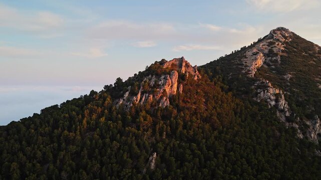 Aerial drone video of a forest-covered mountain peak in Spain at sunrise, with warm sunlight casting golden tones on rocky slopes.