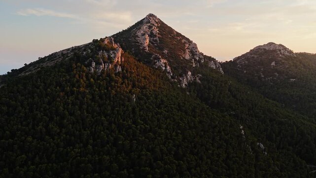 Aerial drone video of a forest-covered mountain peak in Spain at sunrise, with warm sunlight casting golden tones on rocky slopes.