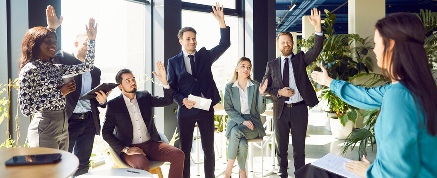 Young confident diverse colleagues raising hands to vote at the conference in meeting room. Business people voting in office during a meeting and listening their female speaker. Banner.