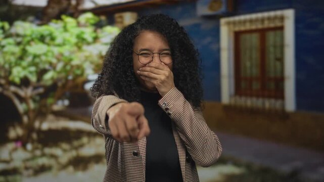 Woman laughing outdoors points playfully at camera with vibrant curly hair in a stylish jacket against a colorful background highlighting cheerful nature.