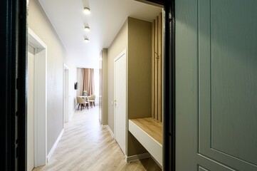 bright hallway with light wood floors leads to a dining area. A built-in bench with wooden slats is near the entrance, framed by a blue-grey door