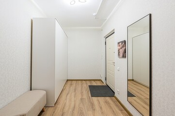 Bright hallway featuring a white wardrobe, a bench, a door with a welcome mat, and a mirror reflecting the scene. Neutral tones and simple decor create a clean aesthetic