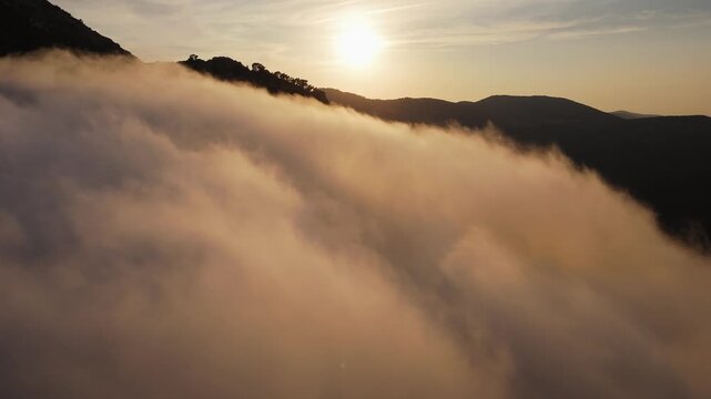 Drone video of sunset casting golden light over a forested Spanish mountain, with thick clouds and mist covering the slopes.