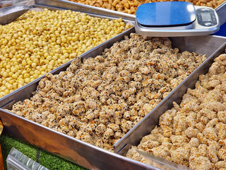 Market display with metal trays of assorted snacks, including sesame-coated nuts, with a digital weighing scale on top.