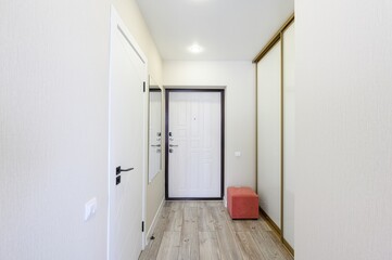 bright hallway features white walls, a white front door with a dark frame, a sliding wardrobe, and a wooden floor. An orange pouf adds a pop of color