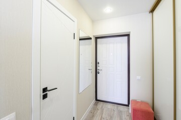 bright hallway features white walls, a white front door with a dark frame, a sliding wardrobe, and a wooden floor. An orange pouf adds a pop of color