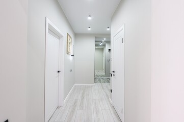 hallway with light gray walls, white doors, and light wood floors. There's a mirror at the end, and recessed lighting illuminates the space