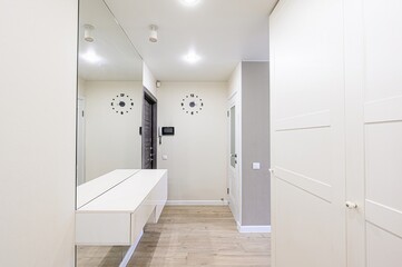 Bright hallway with a mirror, white storage, and wood floors. Two clocks adorn the wall, flanking a dark door