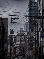Dense urban street scene in South Korea, with tangled overhead power lines, apartment buildings, and moody overcast skies, capturing gritty city life.