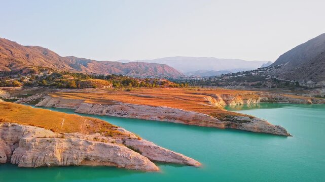 Drone video showing a sharp rocky peninsula surrounded by vibrant turquoise waters of a Spanish lake, bathed in warm golden hour sunlight.
