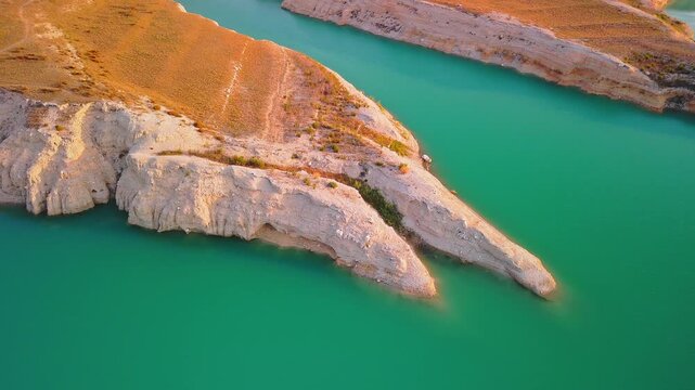 Drone video showing a sharp rocky peninsula surrounded by vibrant turquoise waters of a Spanish lake, bathed in warm golden hour sunlight.