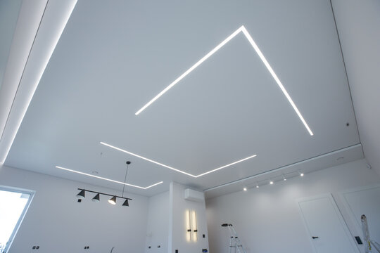 modern room interior with an emphasis on the ceiling. The ceiling is equipped with LED linear lights, creating a geometric pattern. Track lights, a pendant light and wall sconces are also visible.