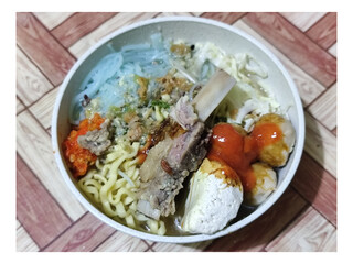 Bakso. A bowl of rib meatballs with yellow noodles, blue vermicelli, shredded cabbage, fried onions and chili sauce on a wooden patterned table. The meatballs look very delicious and delicious.