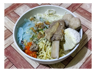 Bakso. A bowl of rib meatballs with yellow noodles, blue vermicelli, shredded cabbage, fried onions and chili sauce on a wooden patterned table. The meatballs look very delicious and delicious.