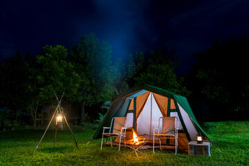 Camping tents on the green grass at night, background with many big trees, vintage cabin tents, antique oil lamps, retro chairs