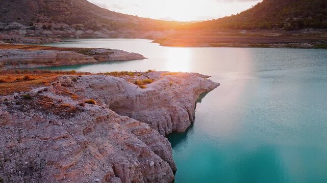 Drone video capturing the rocky lake shore in Spain at sunset, with golden sunlight reflecting on turquoise water and surrounding rugged terrain.