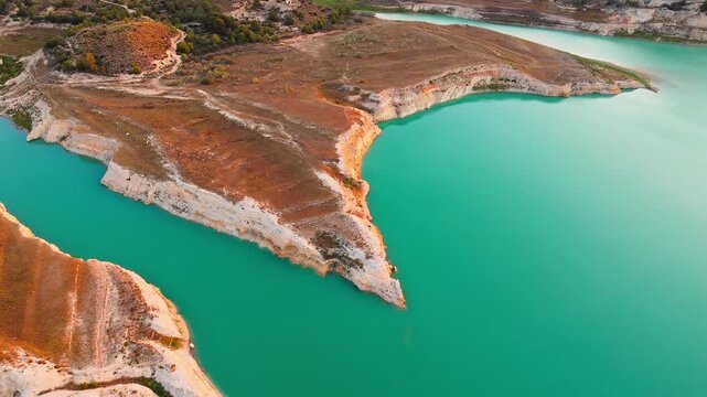 Drone video showing a vivid turquoise lake in Spain with rocky, sunlit shoreline and earthy cliffs creating striking contrast between water and land.