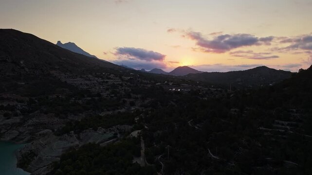 Drone video of a mountain lake in Spain at sunset, with calm turquoise water, rugged hills, and a dramatic sky blending warm and cool tones.