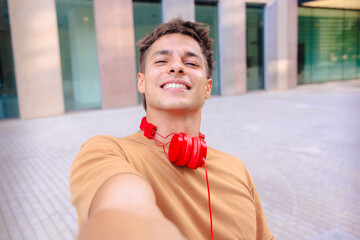 Happy young man wearing a brown t-shirt and red headphones around his neck taking a selfie outdoors...