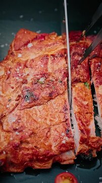 Close-up of juicy grilled pork ribs being sliced with a knife, sprinkled with coarse salt and herbs, served with cherry tomato on black slate.