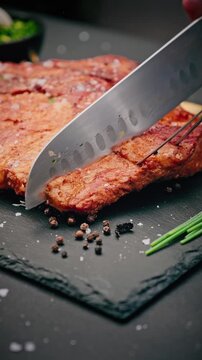 Chef cuts juicy grilled pork steak with sharp knife, garnished with fresh cherry tomatoes and garlic cloves on dark surface.