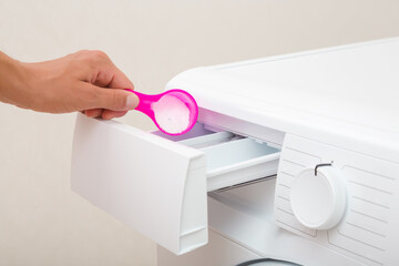 Young adult woman hand holding and pouring pink cup or scoop of white powder in washing machine plastic compartment tray. Clothes care. Light gray background. Closeup. Front view.