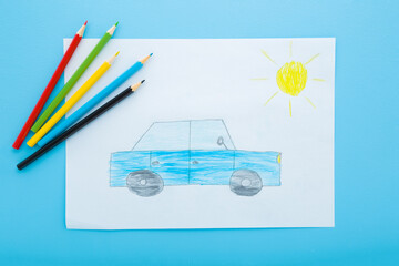Drawn car and sun on white paper. Colorful pencils on light blue table background. Pastel color. Closeup. Little child draw. Top down view.
