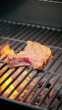 Close-up of raw pork rib cooking over bright orange flames on a barbecue grill with hot charcoal beneath.