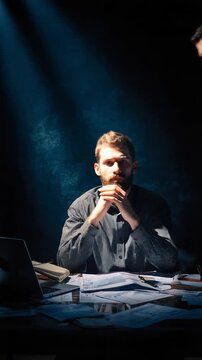 Focused Contemplation: A man sits at a desk under a spotlight, deep in thought, with a determined expression and papers scattered around him.