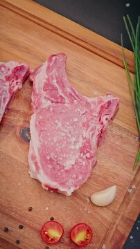 Close-up of chef sprinkling coarse salt on raw pork chops placed on a wooden board with paprika, garlic, and herbs for cooking preparation.