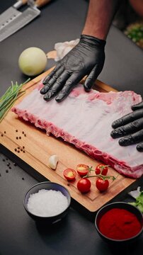 Chef wearing black gloves places raw pork ribs on wooden board with cherry tomatoes, garlic, chives, peppercorns, salt, and paprika nearby.
