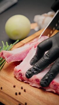 Close-up of chef wearing black gloves slicing raw pork ribs on a wooden board with onion, chives, and peppercorns nearby