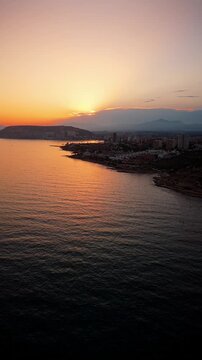 Drone video capturing Spanish coastline and city skyline at sunset, with golden light reflecting on calm sea and rugged shore.