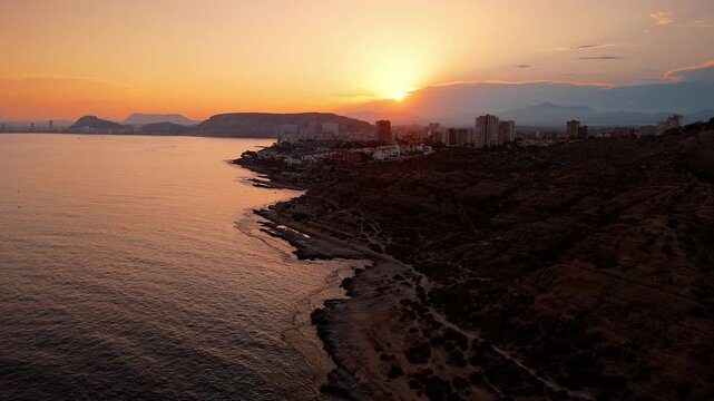 Drone video capturing Spanish coastline and city skyline at sunset, with golden light reflecting on calm sea and rugged shore.