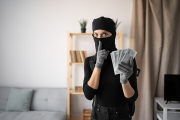 Female thief in black balaclava with cash dollars showing shh silence gesture. Apartment theft....