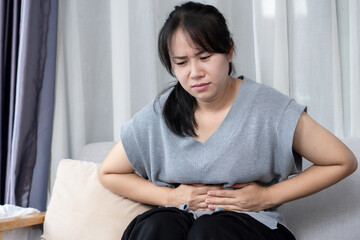 Asian woman experiencing stomach pain while sitting on sofa at home ,signs of indigestion, food...