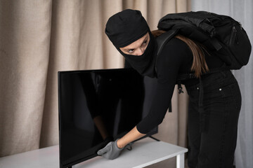 Young Woman burglar in black balaclava stealing tv set from house in daytime