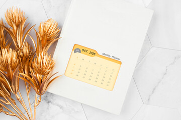 October 2026 calendar on monthly planner book with golden flower on white marble background.