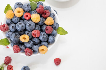 Blueberry and raspberry is tasty, healthy and delicious fruits. Fresh blueberries with red and yellow raspberries with leaves of mint in white bowl. Wild berry in white plate on white background.