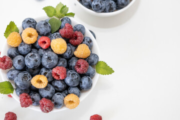 Blueberry and raspberry is tasty, healthy and delicious fruits. Fresh blueberries with red and yellow raspberries with leaves of mint in white bowl. Wild berry in white plate on white background.