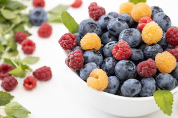 Blueberry and raspberry is tasty, healthy and delicious fruits. Fresh blueberries with red and yellow raspberries with leaves of mint in white bowl. Wild berry in white plate on white background.