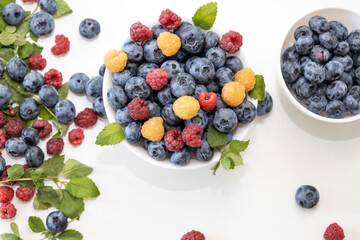 Blueberry and raspberry is tasty, healthy and delicious fruits. Fresh blueberries with red and yellow raspberries with leaves of mint in white bowl. Wild berry in white plate on white background.