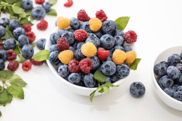 Blueberry and raspberry is tasty, healthy and delicious fruits. Fresh blueberries with red and yellow raspberries with leaves of mint in white bowl. Wild berry in white plate on white background.