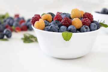 Blueberry and raspberry is tasty, healthy and delicious fruits. Fresh blueberries with red and yellow raspberries with leaves of mint in white bowl. Wild berry in white plate on white background.