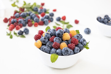 Blueberry and raspberry is tasty, healthy and delicious fruits. Fresh blueberries with red and yellow raspberries with leaves of mint in white bowl. Wild berry in white plate on white background.